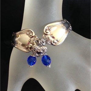 Pode incluir: Pulseira de talheres com cabe&ccedil;as de colher ornamentadas conectadas por uma pe&ccedil;a central decorativa. Duas contas azuis facetadas pendem do centro. A pulseira &eacute; exibida em uma superf&iacute;cie clara.