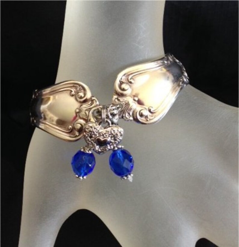 Pode incluir: Pulseira de talheres com cabe&ccedil;as de colher ornamentadas conectadas por uma pe&ccedil;a central decorativa. Duas contas azuis facetadas pendem do centro. A pulseira &eacute; exibida em uma superf&iacute;cie clara.