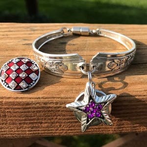 May include: A silver spoon bracelet with a red and white checkered button and a star charm with purple rhinestones.