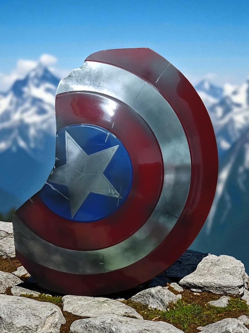 May include: A distressed Captain America shield, featuring a blue center with a silver star, surrounded by red and silver concentric rings. The shield has a battle-worn appearance with visible damage. The background shows a mountain range and blue sky.