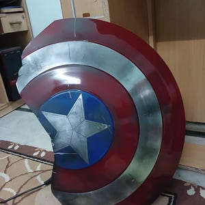 May include: A Captain America shield replica. The shield features a red, white, and blue color scheme with a silver star in the center. The shield has a circular shape with a weathered appearance.
