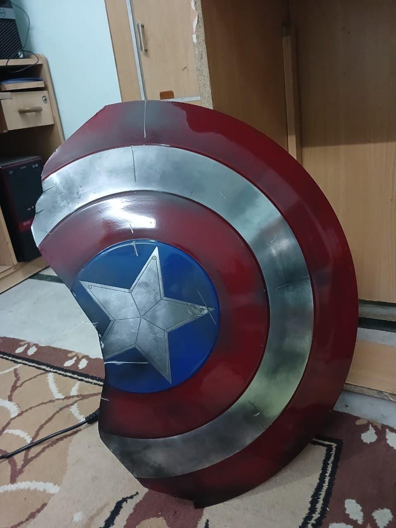 May include: A Captain America shield replica. The shield features a red, white, and blue color scheme with a silver star in the center. The shield has a circular shape with a weathered appearance.