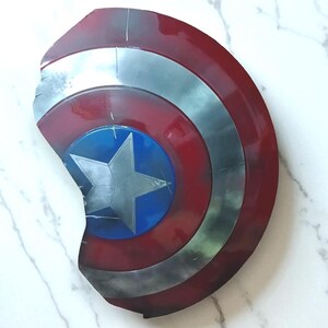 May include: A Captain America shield, featuring a red, white, and blue color scheme. The shield has a circular design with a central blue circle and a silver star. The outer ring is red, and the shield has a distressed, battle-worn appearance.