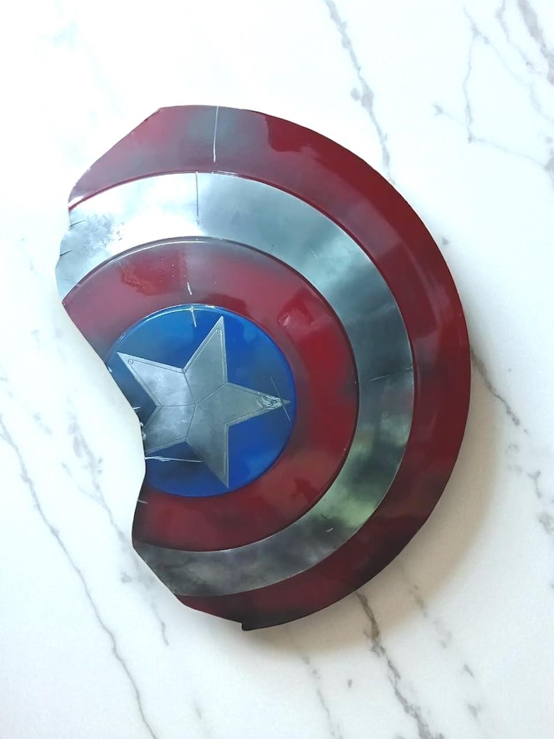 May include: A Captain America shield, featuring a red, white, and blue color scheme. The shield has a circular design with a central blue circle and a silver star. The outer ring is red, and the shield has a distressed, battle-worn appearance.