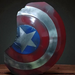May include: A Captain America shield prop, featuring a distressed finish. The shield has a blue center with a white star, surrounded by red and silver concentric circles. The shield is displayed on a wooden surface.