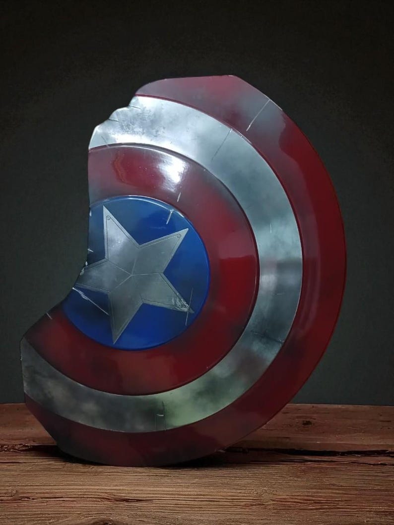 May include: A Captain America shield prop, featuring a distressed finish. The shield has a blue center with a white star, surrounded by red and silver concentric circles. The shield is displayed on a wooden surface.