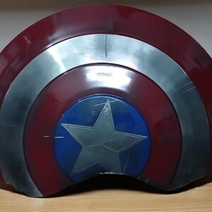 May include: A red, silver, and blue shield with a star emblem. The shield has a circular blue center with a silver star. The outer ring is red, and the middle ring is silver. The shield is likely a prop or collectible.