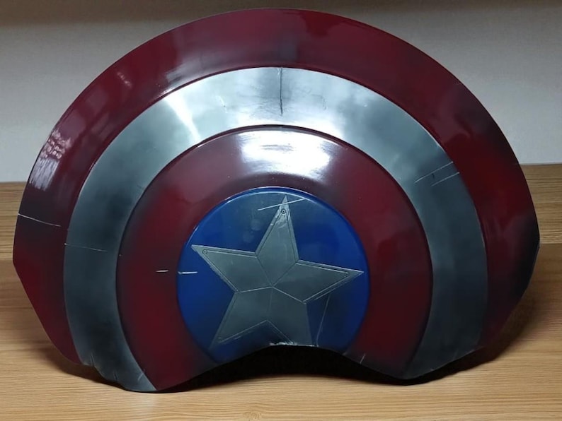 May include: A red, silver, and blue shield with a star emblem. The shield has a circular blue center with a silver star. The outer ring is red, and the middle ring is silver. The shield is likely a prop or collectible.