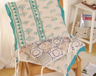 May include: Two decorative chair cushions with a botanical print. The top cushion has a teal border and a repeating pattern of green leaves and triangles. The bottom cushion has a blue and beige floral design. White ties secure the cushions to the chair.