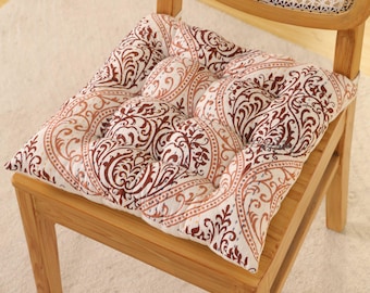 May include: A square chair cushion with a brown and cream paisley pattern. The cushion is placed on a wooden chair with a woven back. The cushion has a tufted design.