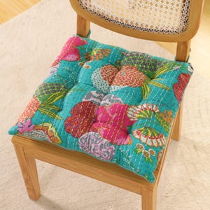 May include: A square, quilted chair cushion with a vibrant floral pattern in shades of teal, pink, orange, and green. The cushion is placed on a wooden chair with a woven back, offering a comfortable and stylish seating option.