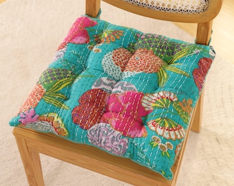 May include: A square, quilted chair cushion with a vibrant floral pattern in shades of teal, pink, orange, and green. The cushion is placed on a wooden chair with a woven back, offering a comfortable and stylish seating option.
