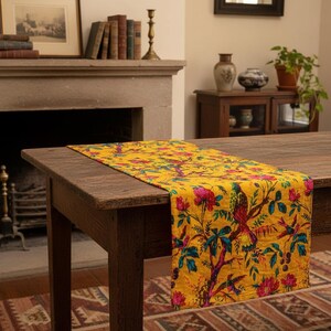 May include: A yellow table runner with a floral and bird pattern. The runner features pink flowers, blue leaves, and birds on a yellow background. The runner is placed on a wooden table.