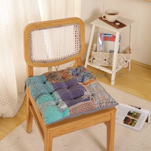 May include: Wooden chair with a woven back and a patchwork cushion. The cushion features a variety of colors and patterns, including teal, purple, and orange. The chair is set on a white rug.