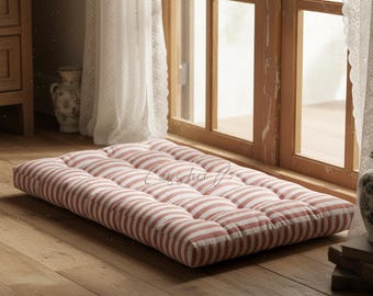May include: A rectangular floor cushion with a red and white striped pattern. The cushion is tufted, with a series of evenly spaced, stitched sections. It rests on a wooden floor near a window.