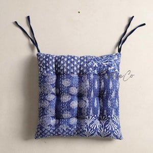 May include: A square seat cushion featuring a patchwork design of blue and white patterns, including concentric circles, leaves, and geometric shapes. The cushion has tufted sections and two dark blue fabric ties at the top corners for securing.