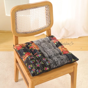 Könnte beinhalten: Ein quadratisches Kissen mit Patchwork-Design, das florale Muster in Rot, Orange und Rosa auf schwarzem Hintergrund aufweist. Das Kissen liegt auf einem Holzstuhl mit gewebter Rückenlehne. Der Text "Cushie Co" ist sichtbar.