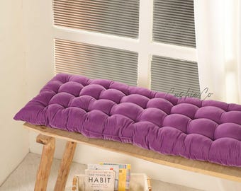 Puede incluir: Un cojín rectangular morado y acolchado descansa sobre un banco de madera. El cojín es de un color morado intenso y vibrante. El banco es de madera clara. El libro "The Power of Habit" es visible.