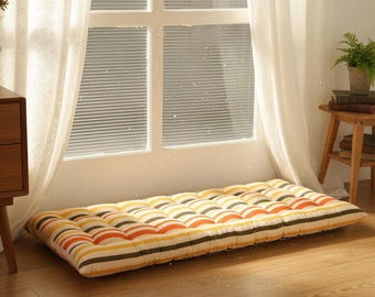 May include: A rectangular floor cushion with a striped pattern in yellow, orange, and black. The cushion is placed on a wooden floor near a window with white curtains. The cushion is approximately 70 inches long and 35 inches wide.