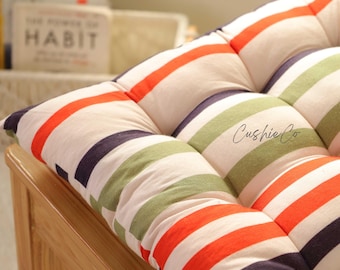 Peut inclure: Coussin rembourré à rayures orange, vert et bleu marine sur fond blanc cassé. Le coussin est matelassé et porte l'inscription "Cushie Co". Il est posé sur une chaise en bois.