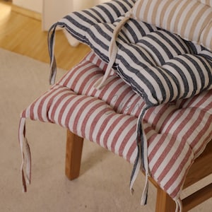 May include: Close-up of three striped seat cushions in red and white, navy and white, and beige and white. The cushions are on a wooden chair and have ties for securing them. The cushions are a home decor item.