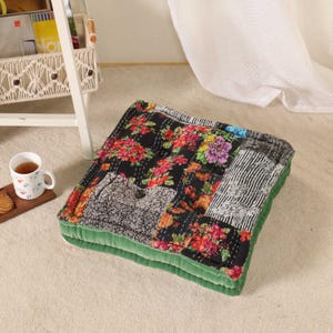 May include: Square floor cushion with a patchwork design featuring floral patterns in red, pink, and purple on a black background. The cushion has a green velvet border and is placed on a light-colored carpet.