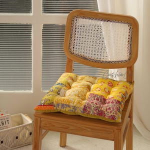 Könnte beinhalten: Holzstuhl mit geflochtener Rückenlehne und gemustertem Kissen. Das Kissen hat gelbe, rote und orangefarbene Stoffe. Der Stuhl steht in der Nähe eines Fensters mit horizontalen Jalousien. Das Wort "Cushie" ist sichtbar.