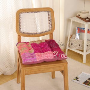May include: A wooden chair with a light-colored cane backrest features a square seat cushion. The cushion is made of patchwork fabric in various shades of pink, magenta, and gold, with visible stitched detailing and a tufted, quilted texture.