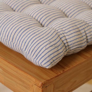 Peut inclure: Gros plan sur un coussin capitonné avec un fond crème et des rayures verticales bleues. Le coussin repose sur une surface en bois brun clair. Le design du coussin est un motif classique et simple.