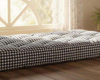 May include: A black and white checkered futon mattress. The mattress is rectangular and tufted, with a grid pattern. It is placed on a wooden floor near a window with a sheer curtain.