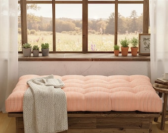 May include: A peach-colored, tufted cushion sits atop a rustic wooden bench. A light gray knitted throw blanket is draped over the cushion. The bench is positioned in front of a large window with a view of a field and several potted plants.