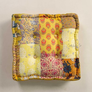 May include: A square floor cushion with a patchwork design. The cushion features various yellow fabrics with floral and paisley patterns, stitched together. It has button tufting and a quilted border. The brand name "Cushie Co" is visible on the cushion.
