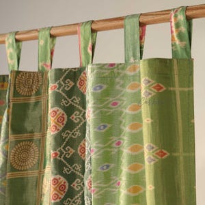 May include: A collection of green and patterned fabric curtains hanging from a wooden rod. The curtains feature various designs, including geometric and floral patterns in shades of green, yellow, and red. The fabric has a silky sheen.