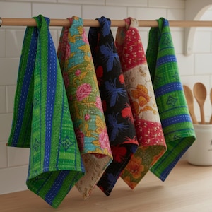 Può includere: Una collezione di cinque strofinacci da cucina colorati appesi a un'asta di legno. Gli strofinacci presentano vari motivi, tra cui disegni floreali, strisce e forme geometriche. La tavolozza dei colori include verde, blu, rosso e nero.