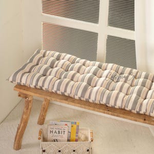 May include: A long, tufted bench cushion with a striped pattern in beige, grey, and white. The cushion is placed on a rustic wooden bench. A small basket with books sits below the bench near a window.