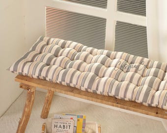May include: A long, tufted bench cushion with a striped pattern in beige, gray, and white. The cushion is placed on a rustic wooden bench. A small basket with books sits below the bench near a window.