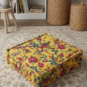 May include: Square floor cushion featuring a yellow background with a floral and bird design in pink, purple, and blue. The cushion is quilted and appears thick and padded. Ideal for a living room.