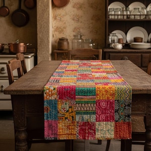 May include: A colorful patchwork table runner with a variety of patterns and colors, including pink, yellow, and blue, rests on a dark wooden table. The runner is made of squares of fabric sewn together. The scene is set in a rustic kitchen.