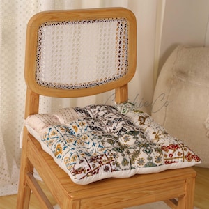 Puede incluir: Una silla de madera con respaldo tejido y un cojín estampado. El cojín presenta un diseño de patchwork con varios colores, incluyendo azul, marrón y blanco. La silla es de madera clara.