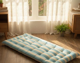 May include: A rectangular floor cushion with a light blue and white striped pattern. The cushion is made of a soft, quilted material and is designed for comfort. It is placed on a wooden floor in a well-lit room.
