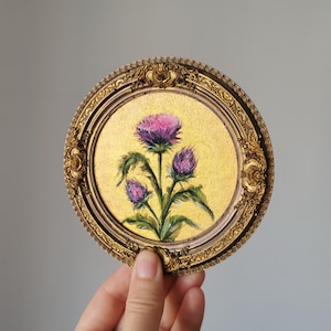 Puede incluir: Pintura redonda enmarcada en oro que representa un cardo con flores moradas y hojas verdes. El fondo es dorado. El marco dorado está adornado con detalles. La pintura está sostenida por una mano.