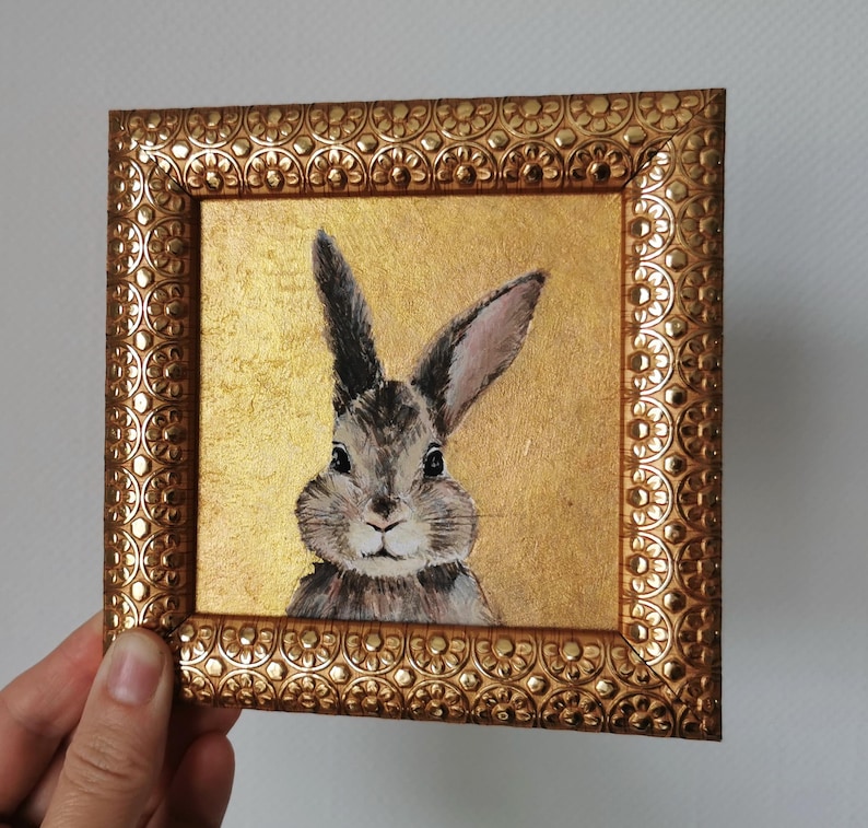 Hand holding framed acrylic bunny painting with golden background.