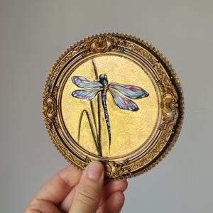Könnte beinhalten: Ein rundes, goldfarbenes Dekorationsobjekt mit einer detaillierten Malerei einer Libelle. Die Libelle hat schillernde blaue, violette und grüne Flügel und einen schlanken Körper. Es ist mit einem verzierten, antiken Goldrahmen eingefasst.