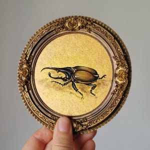 Może przedstawiać: Okrągła, ozdobna, złota rama otacza złote, teksturowane tło z szczegółowym obrazem brązowego i czarnego chrząszcza. Rama jest trzymana przez dłoń.