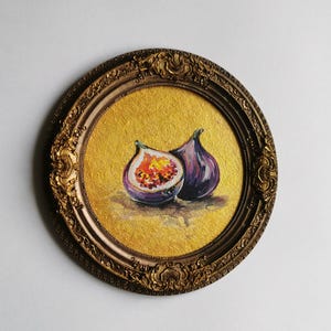Könnte beinhalten: Gerahmtes Gemälde mit zwei Feigen. Eine Feige ist halbiert und zeigt ein farbenfrohes Inneres aus Rot, Orange und Gelb. Die andere Feige ist ganz. Der Hintergrund ist goldfarben, der Rahmen ebenfalls.
