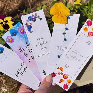 Könnte beinhalten: Eine Sammlung floraler Lesezeichen mit verschiedenen Designs. Jedes Lesezeichen zeigt gepresste Blumen und handgeschriebenen Text, darunter Sätze wie "Neues Kapitel". Die Lesezeichen werden vor einem Hintergrund aus Blumen gehalten.