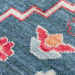 K&ouml;nnte beinhalten: Nahaufnahme eines blauen Teppichs mit strukturierter Oberfl&auml;che. Der Teppich weist ein sich wiederholendes Muster aus wei&szlig;en, roten und orangefarbenen Blumen- und geometrischen Designs auf. Das Design des Teppichs umfasst ein Zickzackmuster.