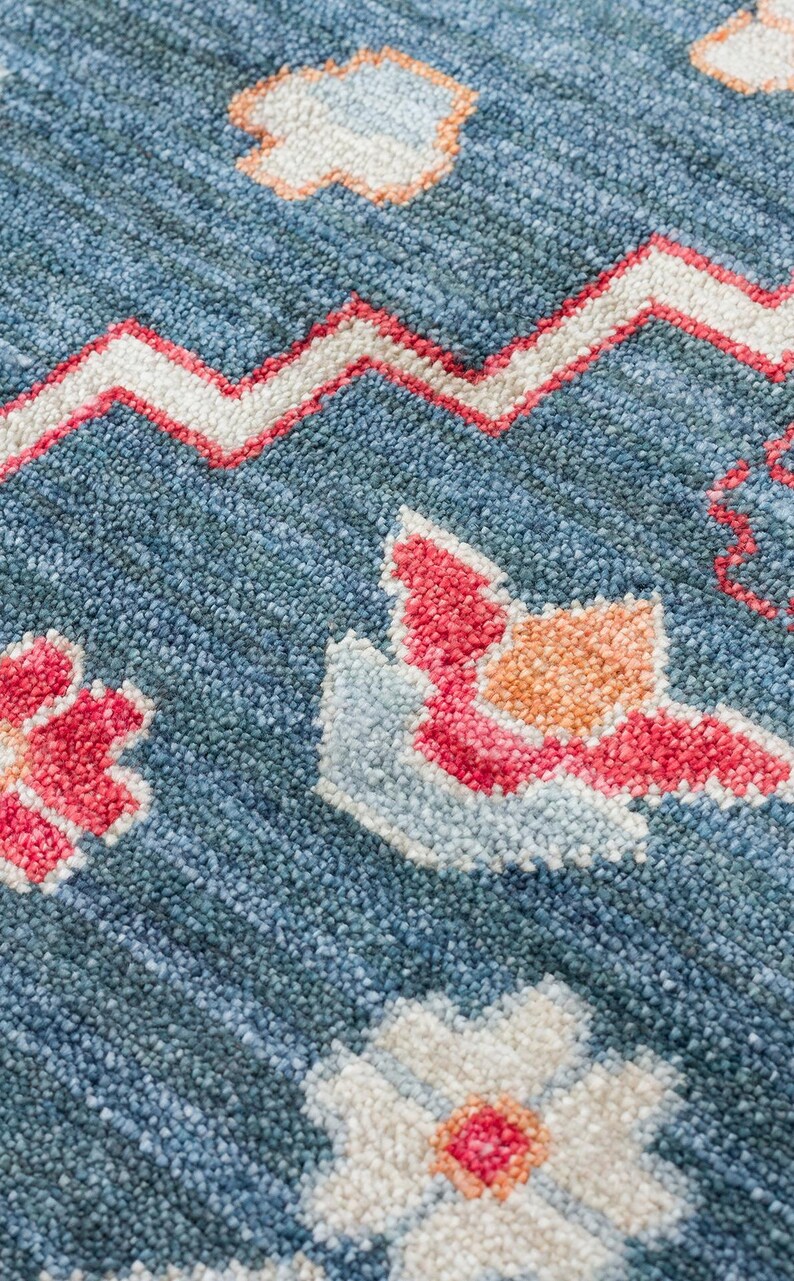 K&ouml;nnte beinhalten: Nahaufnahme eines blauen Teppichs mit strukturierter Oberfl&auml;che. Der Teppich weist ein sich wiederholendes Muster aus wei&szlig;en, roten und orangefarbenen Blumen- und geometrischen Designs auf. Das Design des Teppichs umfasst ein Zickzackmuster.