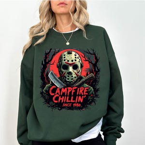 May include: Dark green sweatshirt featuring a graphic of a hockey mask, a knife, and the text "Campfire Chillin' Since 1980." The design includes a red moon and silhouettes of trees. The sweatshirt is a crew neck style.