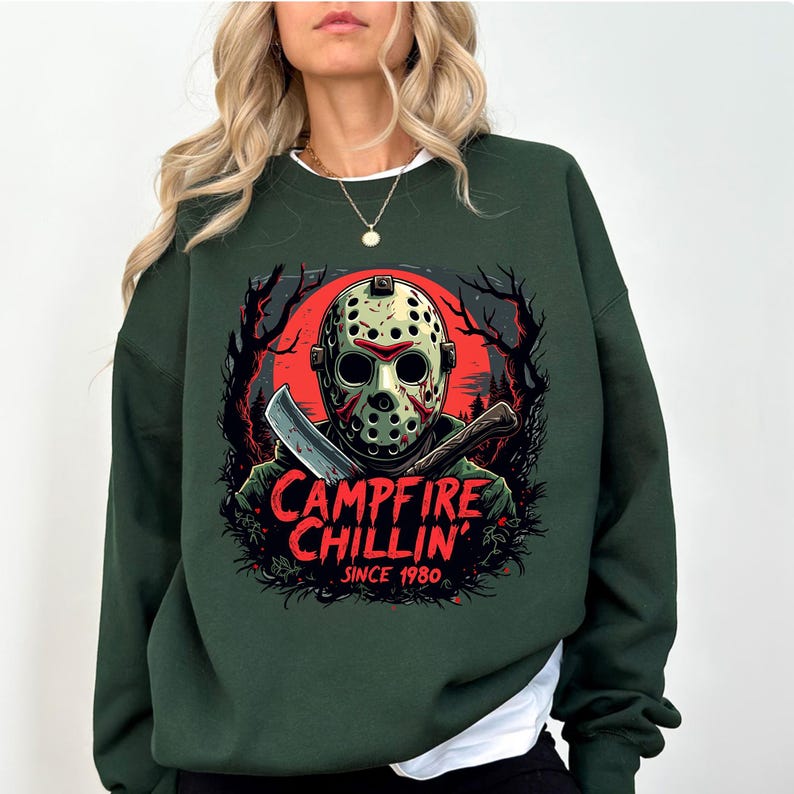 May include: Dark green sweatshirt featuring a graphic of a hockey mask, a knife, and the text "Campfire Chillin' Since 1980." The design includes a red moon and silhouettes of trees. The sweatshirt is a crew neck style.